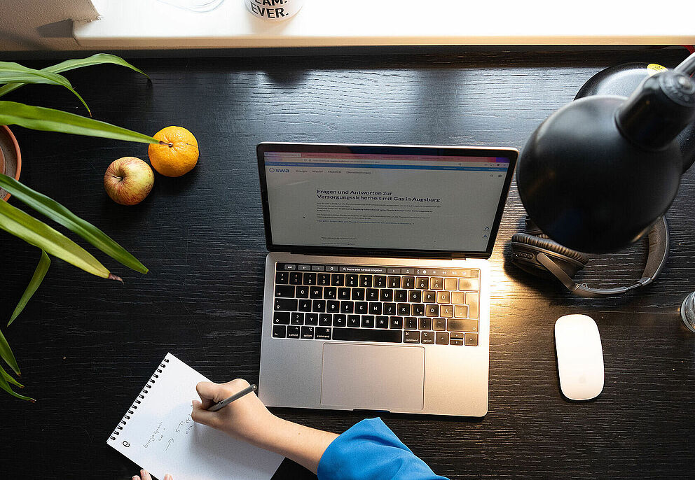 Person sitzt an einem Schreibtisch und schreibt in ein Notizbuch. Auf dem Tisch ist ein Laptop eine Lampe und Obst