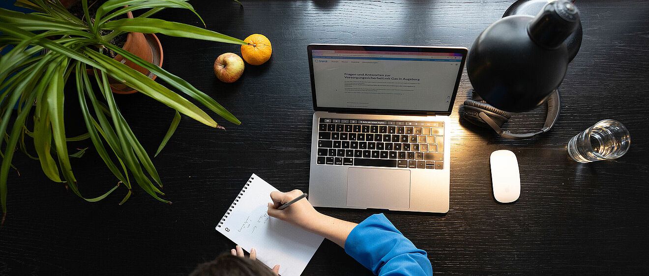 Person sitzt an einem Schreibtisch und schreibt in ein Notizbuch. Auf dem Tisch ist ein Laptop eine Lampe und Obst