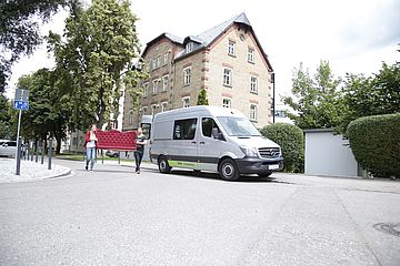 Swa Carsharing Transporter steht vor einem Haus. Daneben tragen zwei Personen ein Sofa. - Ein Klick auf das Bild führt zur vergrößerten Fotoansicht. 