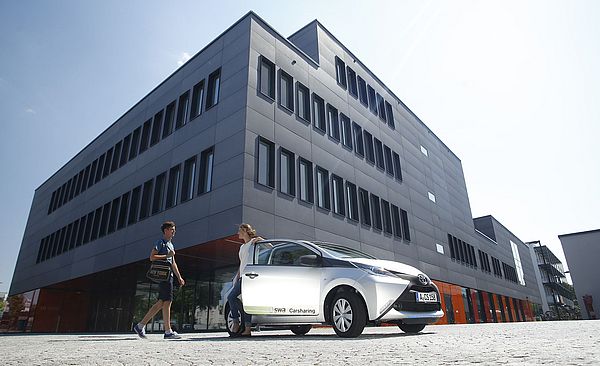 Swa Carsharing Auto vor einem großen Gebäude. Am Auto unterhalten sich zwei Personen. 