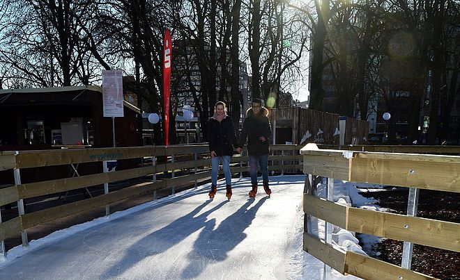 Eine junge Frau und ein junger Mann laufen händchenhaltend Schlittschuh auf einer Eisbahn am Augsburger Königsplatz.