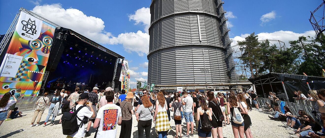 Menschen stehen auf dem Gaswerksgelände. Im Hintergrund ist eine Bühne und der große Gasbehälter. Weiterverlinkung auf swa und Modular Partnerschaft Newsartikel