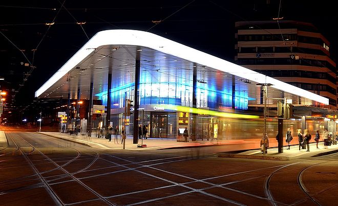 Im Vordergrund kreuzen sich die Schienen der Straßenbahn auf dem beleuchteten Platz. In der Bildmitte befindet sich die moderne, nachts hell beleuchtete Haltestelle Königsplatz in Augsburg, deren geschwungenes Dach und Glasfront bläulich leuchten.
