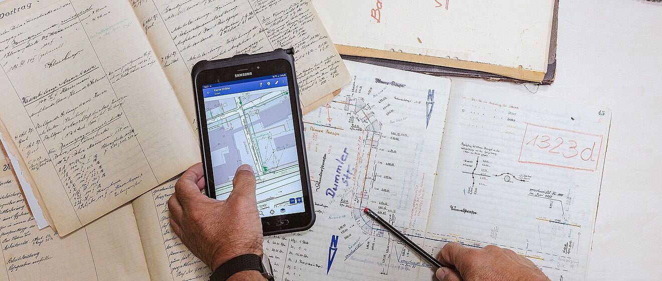Hand hält ein Tablet mit Zeichnungen darunter ist ein Tisch mit vollen Notizblöcken. Link zum Öffnen eines Pop-Ups mit Informationen zu Vermessungen von Leitungen und Anlagen bis zum 3D-Scanning