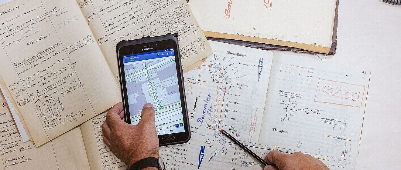 Hand hält ein Tablet mit Zeichnungen darunter ist ein Tisch mit vollen Notizblöcken. Link zum Öffnen eines Pop-Ups mit Informationen zu Vermessungen von Leitungen und Anlagen bis zum 3D-Scanning