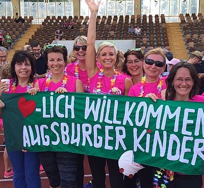 Teilnehmerinnen des Frauenlaufs stehen nebeneinander und halten ein Banner mit der Aufschrift "Herzlich Willkommen Augsburger Kinder" in die Kamera.