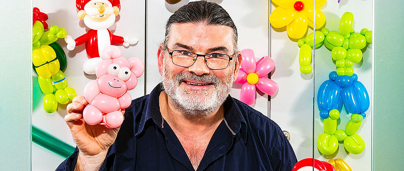 Ein Mann schaut lächelnd in die Kamera und hält eine Luftballon-Figur in seiner Hand. Im Hintergrund hängen weitere solcher Figuren an einer Wand. 