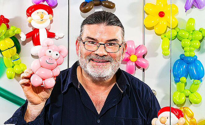 Ein Mann schaut lächelnd in die Kamera und hält eine Luftballon-Figur in seiner Hand. Im Hintergrund hängen weitere solcher Figuren an einer Wand. 