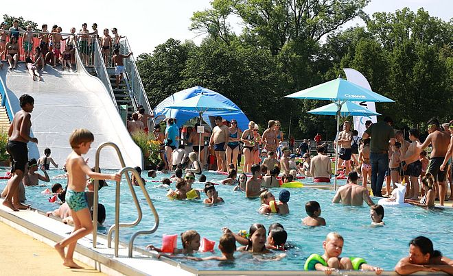 Viele Erwachsene und Kinder im Schwimmbad. Links im Bild eine Wasserrutsche.