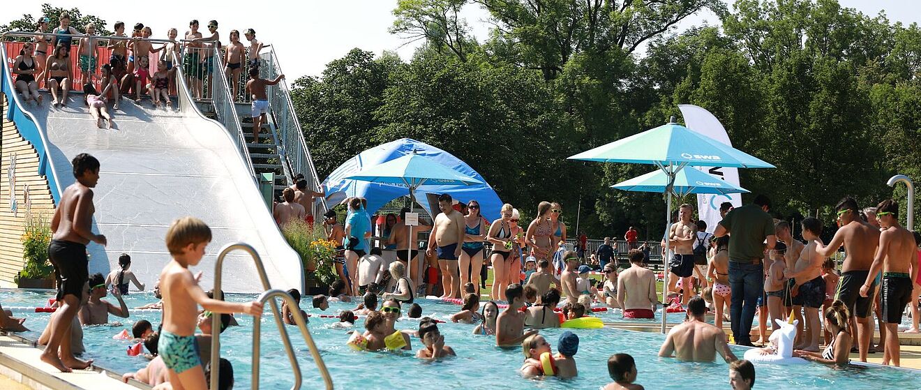 Viele Erwachsene und Kinder im Schwimmbad. Links im Bild eine Wasserrutsche.