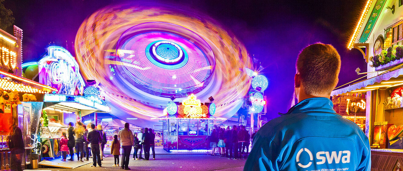 Mann mit swa Jacke auf der Augsburger Plärrer. Im Hintergrund sind Gäste und verschiedene Stände und Fahrattraktionen