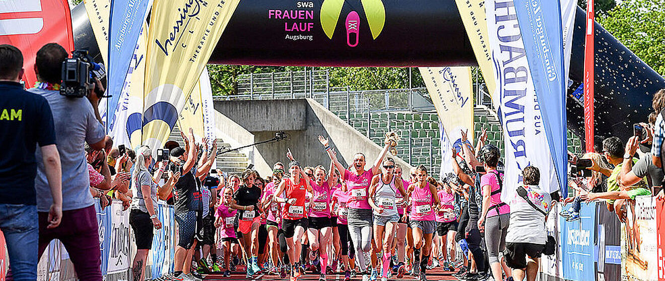 Zentral laufen mehrere Frauen in Sportkleidung jubelnd durch den Zieleinlauf. Auf der linken und rechten Seite befinden sich Zuschauer.