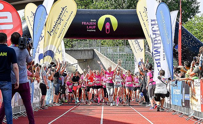 Zentral laufen mehrere Frauen in Sportkleidung jubelnd durch den Zieleinlauf. Auf der linken und rechten Seite befinden sich Zuschauer.