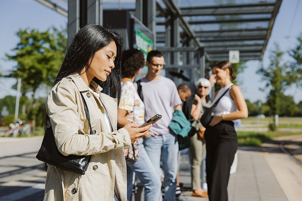Frau steht mit Handy in der Hand an einer Haltestelle