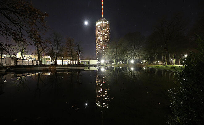 Der erleuchtete Augsburger Hotelturm bei Nacht, der sich im Wasser spiegelt, aus der Sicht des Wittelsbacher Parks.