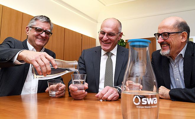 Der swa Geschäftsführer Alfred Müllner, der Oberbürgermeister Dr. Kurt Gibl sowie ein weiterer Mann sitzen lächelnd an einem Tisch und schenken sich ein swa Trinkwasser in Gläsern ein.