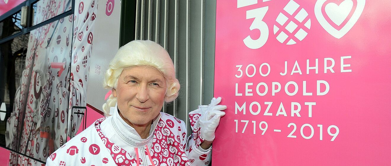 Ein Mann mit pink-weißer Kleidung und einer Mozart-Perücke steht neben einem pinken Schild mit der Aufschrift "Leo300 300 Jahre Leopold Mozart 1719 - 2019""