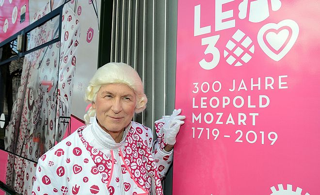 Ein Mann mit pink-weißer Kleidung und einer Mozart-Perücke steht neben einem pinken Schild mit der Aufschrift "Leo300 300 Jahre Leopold Mozart 1719 - 2019""