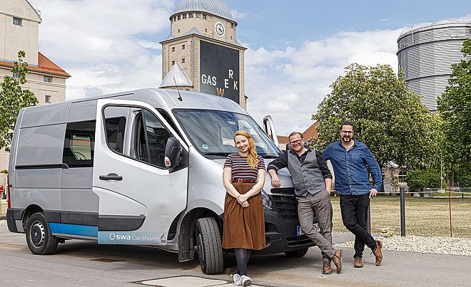 Swa Carsharing Transporter mit geöffneten Vordertüren. Davor eine Frau und zwei Männer, die sich am auto anlehnen. Im Hintergrund die Gaswerke.
