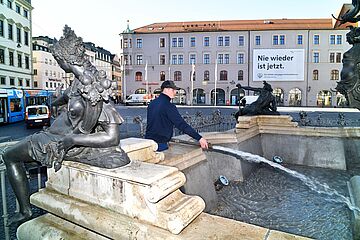 Mann mit Kappe befüllt den Augustusbrunnen am Rathausplatz in Augsburg mit Wasser. Im Hintergrund ist links ein swa Auto und eine Straßenbahn