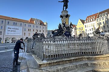 Mann mit Kappe reinigt den Augustbrunnen beim Rathausplatz in Augsburg mit einem Hochdruckreiniger