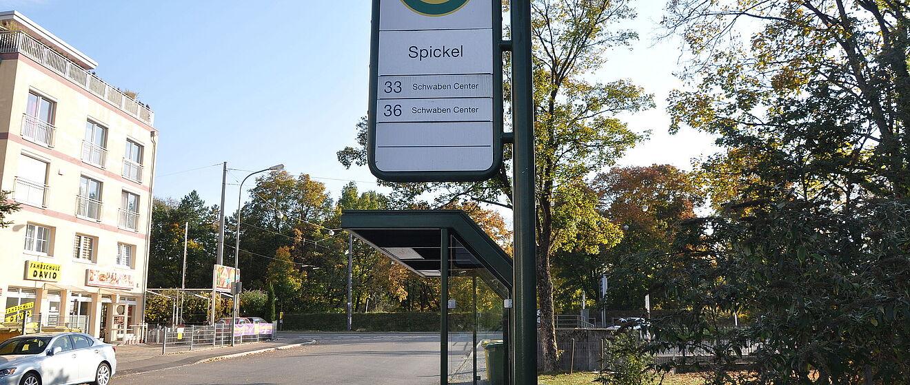 Im Vordergrund ist die Haltestelle mit dem Namen "Spickel". Im Hintergrund befindet sich eine Reihe Bäumen.