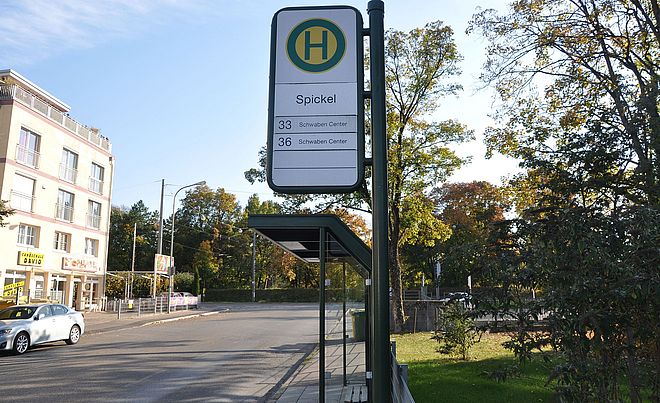 Im Vordergrund ist die Haltestelle mit dem Namen "Spickel". Im Hintergrund befindet sich eine Reihe Bäumen.