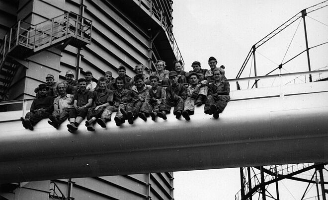 Arbeiter sitzen auf einer Röhre auf der Außenfassade des Gaswerks. Weiterverlinkung auf Industriedenkmal Newsartikel