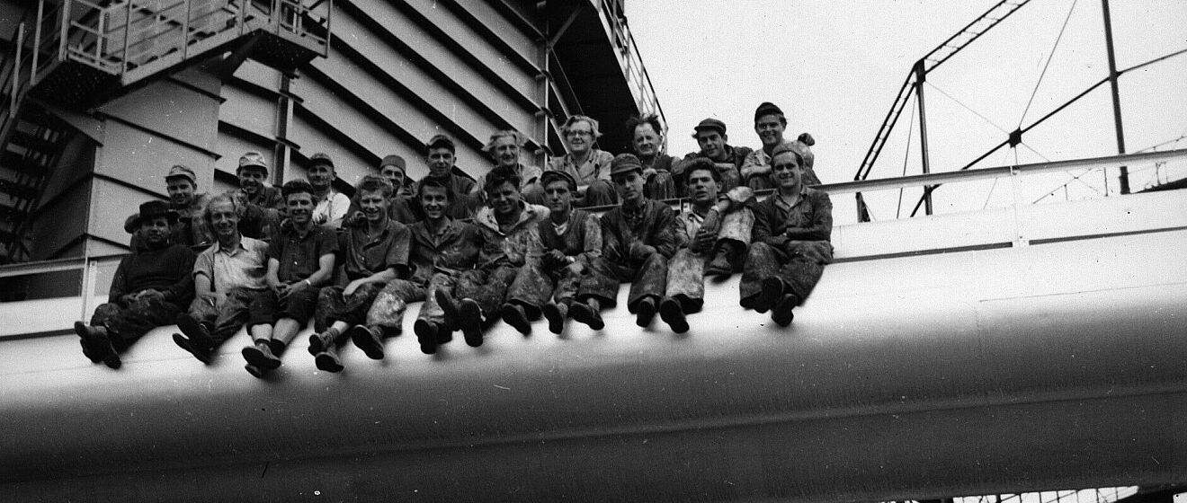 Arbeiter sitzen auf einer Röhre auf der Außenfassade des Gaswerks. Weiterverlinkung auf Industriedenkmal Newsartikel