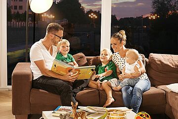 Familie, die gemütlich zusammen auf der Couch sitzt und ein Kinderbuch liest. - Ein Klick auf das Bild führt zur vergrößerten Fotoansicht.