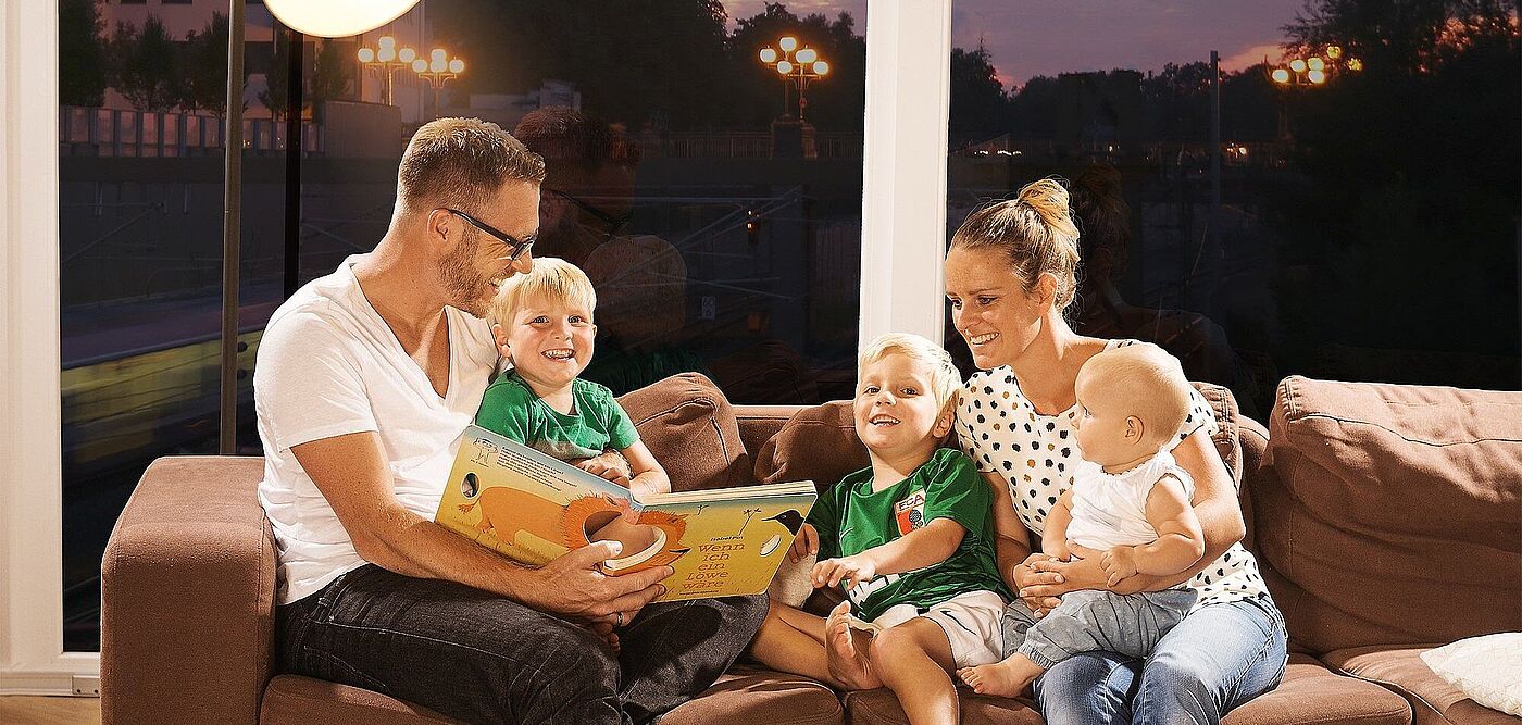 Ein lachender Mann mit einem Kleinkind und einem Buch in der Hand sitzt neben einem Kind und einer lachenden Frau mit einem Baby in der Hand auf einer Couch