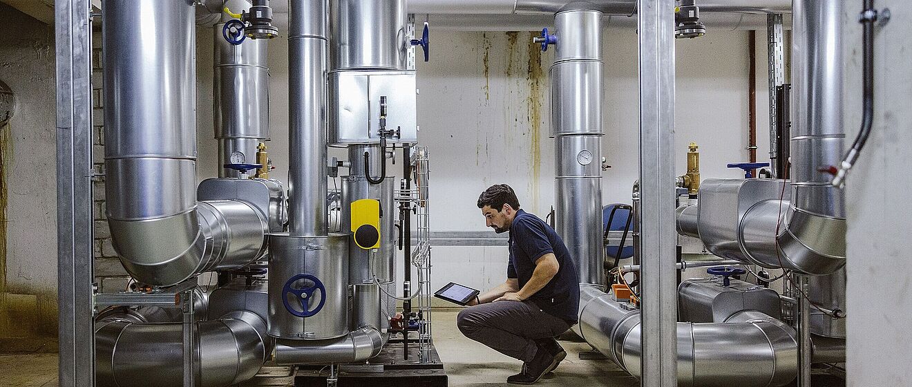 Mann in der Hocke mit einem Tablet. Vor ihm sind technische Anlagen. Link zu einem Pop-Up mit Informationen zu Technischen Energie- und Gebäudemanagement