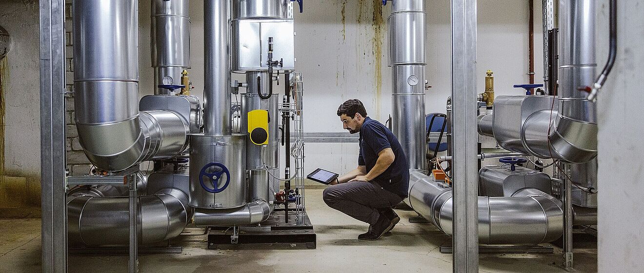 Mann in der Hocke mit einem Tablet. Vor ihm sind technische Anlagen.
