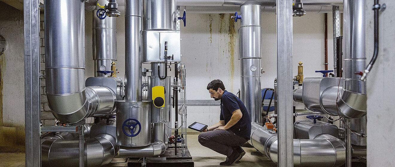 Mann in der Hocke mit einem Tablet. Vor ihm sind technische Anlagen.