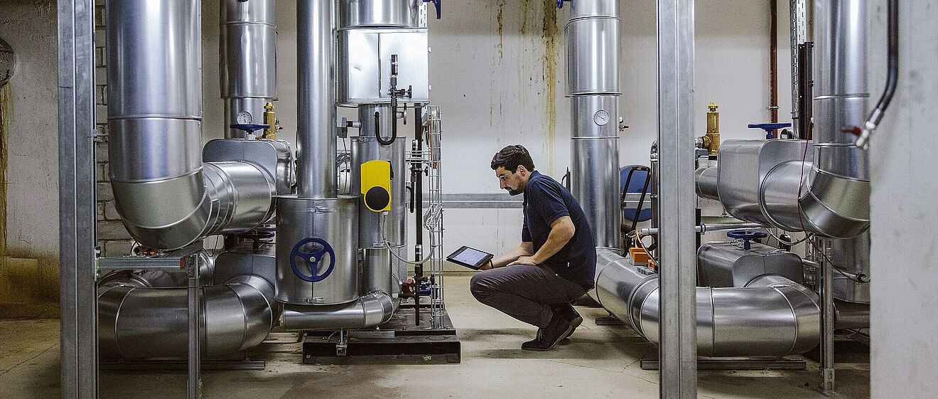 Mann in der Hocke mit einem Tablet. Vor ihm sind technische Anlagen. Link zu einem Pop-Up mit Informationen zu Technischen Energie- und Gebäudemanagement