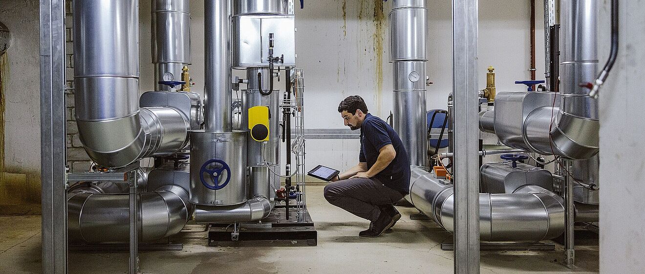 Mann in der Hocke mit einem Tablet. Vor ihm sind technische Anlagen.