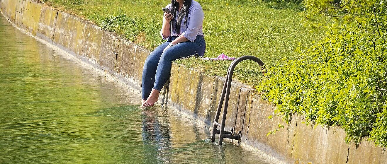 Frau die am Rand der Badestelle am Proviantbach sitzt. Ihre Füße hängen ins Wasser und sie hört mit Kopfhörern Musik.