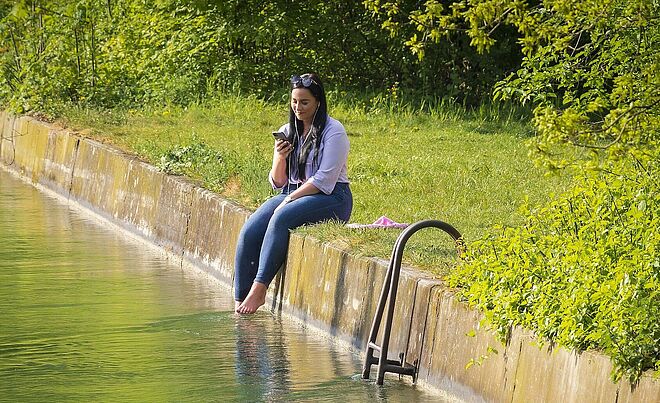 Frau die am Rand der Badestelle am Proviantbach sitzt. Ihre Füße hängen ins Wasser und sie hört mit Kopfhörern Musik.