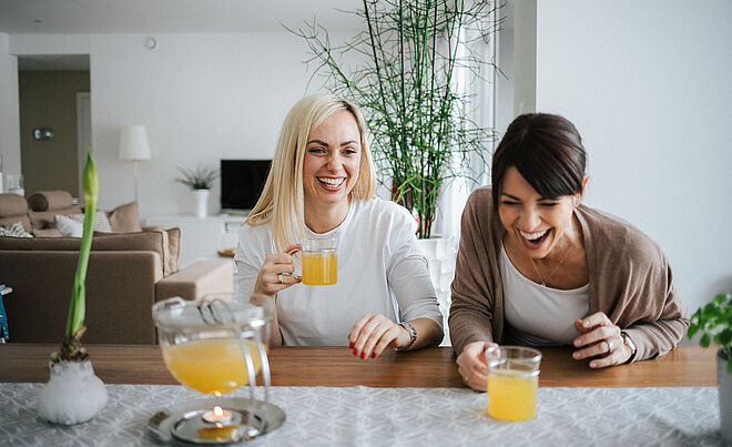 Zwei Bloggerinnen namens Karin und Maja sitzen gemeinsam an einem Tisch und lachen. Dabei halten die beiden Frauen ein Teeglas in der Hand.