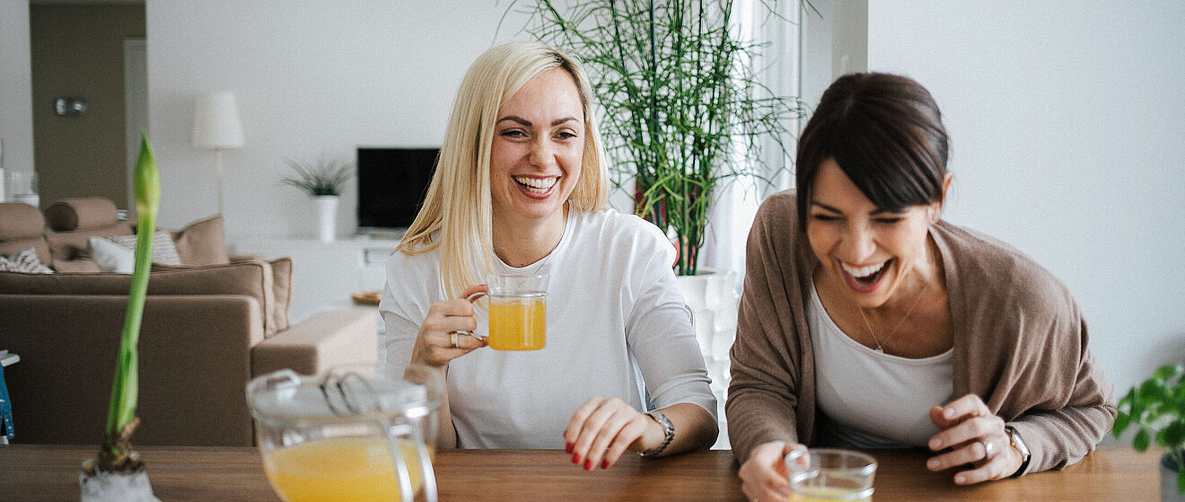 Zwei Bloggerinnen namens Karin und Maja sitzen gemeinsam an einem Tisch und lachen. Dabei halten die beiden Frauen ein Teeglas in der Hand.