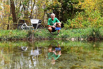 Mann kniet an Brunnenbach.  - Ein Klick auf das Bild führt zur vergrößerten Fotoansicht.