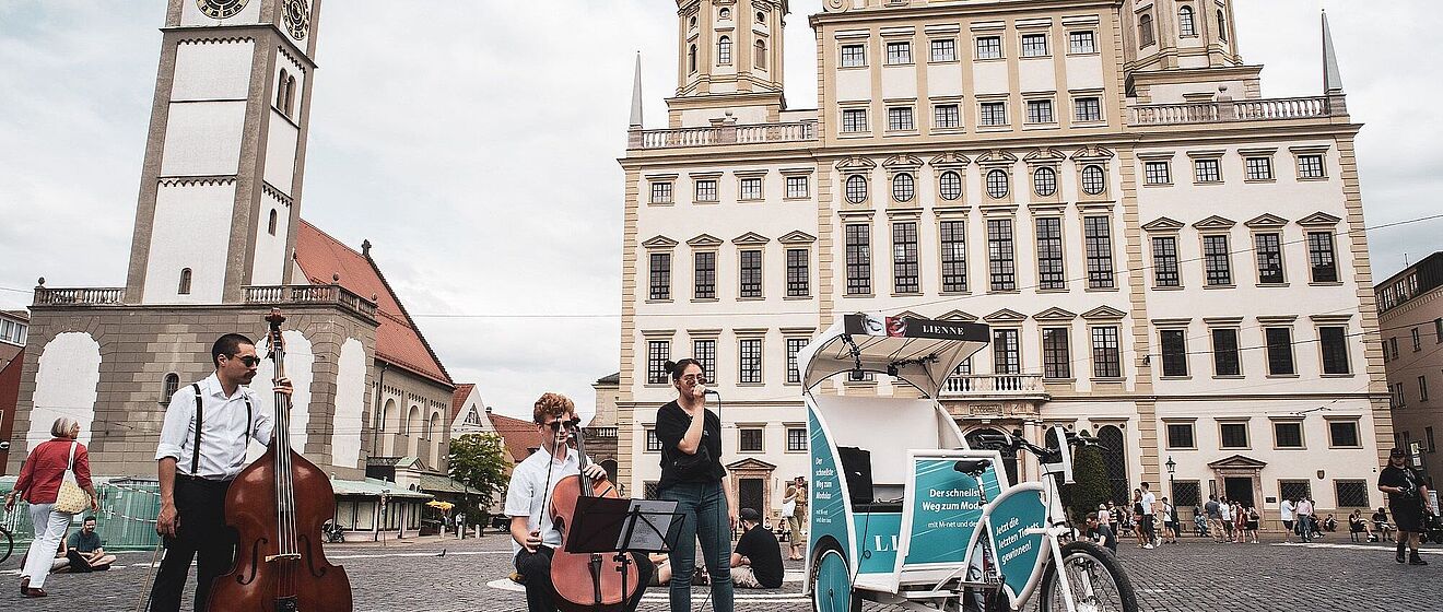 Eine Gruppe von drei Menschen mit Kontrabass und Mikrofon stehen neben einer Fahrradrikscha. Im Hintergrund ist das Augsburger Rathaus zu sehen.