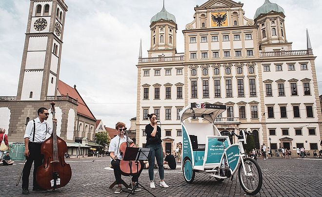 Eine Gruppe von drei Menschen mit Kontrabass und Mikrofon stehen neben einer Fahrradrikscha. Im Hintergrund ist das Augsburger Rathaus zu sehen.
