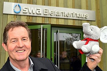 Der Mann im Vordergrund ist Walter Casazza, der ehemalige Geschäftsführer der Stadtwerke Augsburg (swa). Er hält einen kleinen Plüsch-Elefanten in der Hand und steht vor dem neuen swa Elefantenhaus im Augsburger Zoo. Ein Klick auf das Bild führt zu einer größeren Ansicht.
