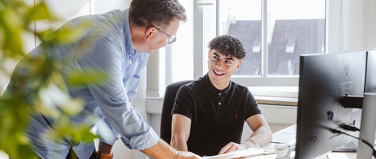 Zwei Kollegen unterhalten sich im Büro an einem Schreibtisch vor Monitoren