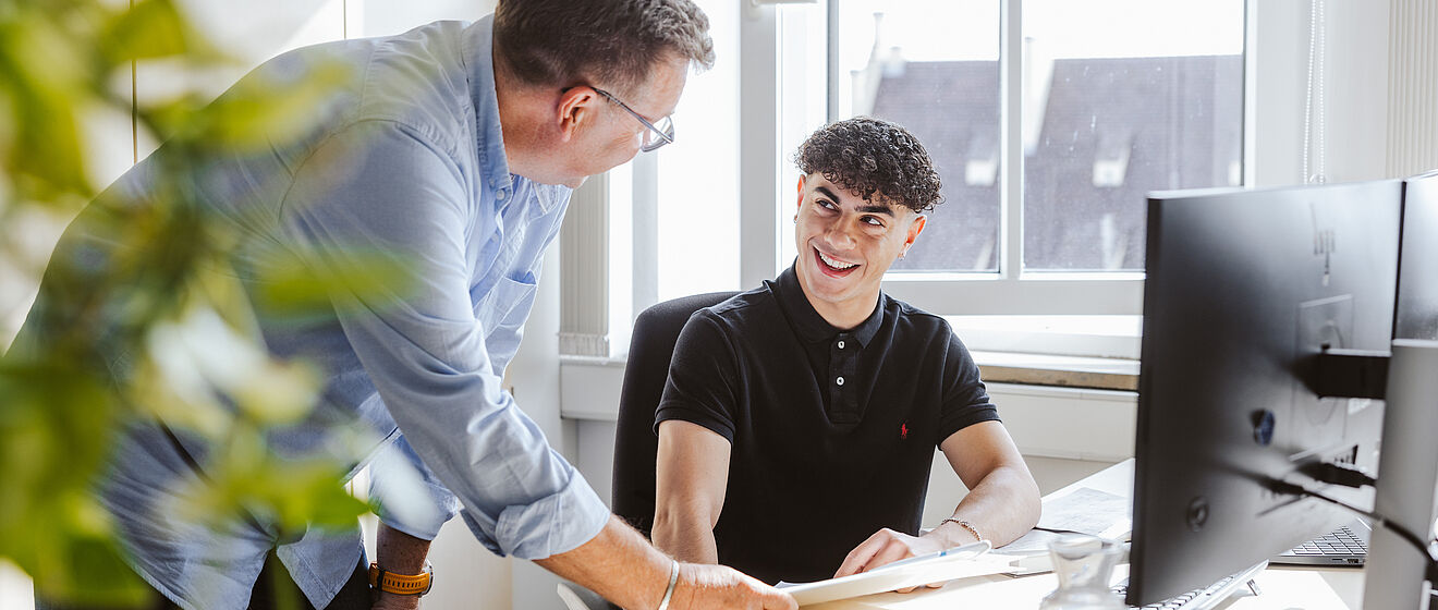 Zwei Kollegen unterhalten sich im Büro an einem Schreibtisch vor Monitoren