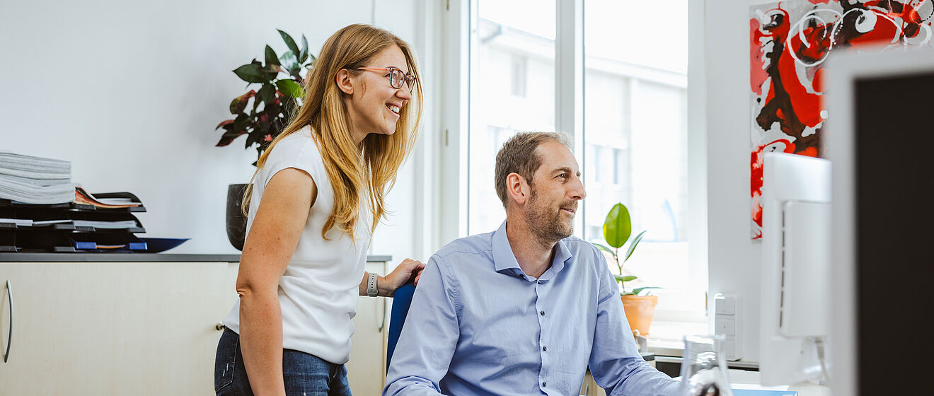 Ein Mann sitzt an einem Schreibtisch und schaut zusammen mit einer hinter ihm stehenden Frau in einen Monitor