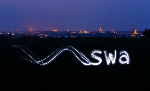 Das Bild zeigt eine nächtliche Panoramaaufnahme der Stadt Augsburg mit leuchtenden Stadtsilhouetten in der Ferne. Im Vordergrund, auf einem dunklen Feld, wurden durch die Technik des Lightpainting die Logos der Stadtwerke Augsburg (swa) mit Licht in die Luft gemalt, was einen kreativen visuellen Effekt erzeugt.