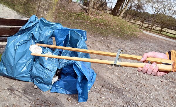 Eine Hand hält eine Greifzange, mit dem ein Stück Müll aufgehoben wird. Dahinter sieht man mehrere Müllbeutel und Bäume.