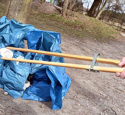 Eine Hand hält eine Greifzange, mit dem ein Stück Müll aufgehoben wird. Dahinter sieht man mehrere Müllbeutel und Bäume.